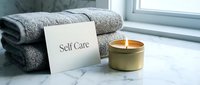 Soft gray towels stacked on marble surface with a lit candle and self care card, creating a serene spa atmosphere for wellness and relaxation.