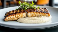 Grilled salmon fillet with crispy charred exterior served on creamy cauliflower puree, garnished with fresh microgreens on white plate against dark background.