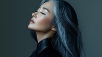 Young Asian woman with long dark blue hair in profile view against dark background, eyes closed in serene expression, dramatic studio lighting creates moody atmosphere.