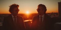 Two professional businessmen in suits having conversation during golden hour sunset with warm lighting and city skyline background for corporate meetings.