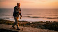 Senior man with walking stick enjoying peaceful sunset walk along coastal wooden boardwalk overlooking ocean waves and horizon during golden hour.