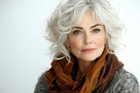 Confident mature woman with silver wavy hair wearing cozy knit sweater and orange scarf smiling warmly against light background, representing aging gracefully and natural beauty.