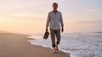 Middle-aged man with dark hair and light skin tone, wearing casual button-up shirt and pants, walking barefoot along sandy beach at sunset carrying shoes in hand, ocean waves in background.