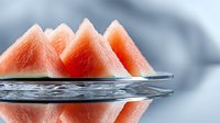 Fresh triangular watermelon slices arranged on glass plate with water droplets, vibrant coral pink flesh against soft gray background, macro food photography.