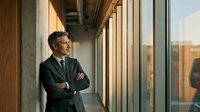 Confident middle-aged Caucasian businessman in dark suit standing with crossed arms in modern office corridor looking through windows at city skyline.