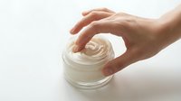 Caucasian hand holding open glass jar of white cosmetic cream or moisturizer on white background. Skincare product application and beauty routine concept.
