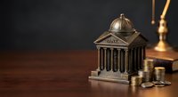 Bronze miniature bank building with classical columns and dome surrounded by stacked coins on wooden surface, symbolizing financial security and savings growth.