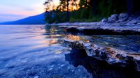 Tranquil lake shore at dusk with rocky outcrop in foreground, silhouetted trees and mountains in background, golden sunset reflections on calm water.