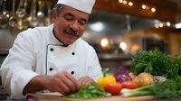 Hispanic senior chef in white uniform carefully chopping fresh vegetables in professional kitchen. Colorful produce and herbs on wooden cutting board with soft lighting.