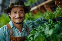 Asian male farmer in hat and overalls smiling while tending to organic vegetables in greenhouse garden, representing sustainable agriculture and local farming.