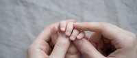 Adult hands gently holding tiny newborn baby fingers in tender moment of connection and care on soft gray background.