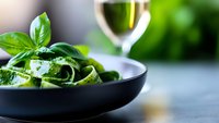 Fresh green zucchini ribbons with basil leaves in black ceramic bowl, white wine glass in background. Selective focus on vegetables with blurred backdrop.