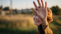 Young woman with red hair waving hand in warm sunset light, showing friendly gesture against blurred natural background.