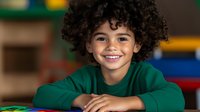 Cheerful mixed race child with curly hair wearing green sweater smiling brightly at camera in classroom setting. Natural authentic expression shows joy and confidence.