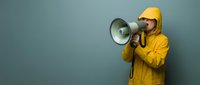 Young man in yellow hoodie and cap shouting into megaphone against blue wall background for communication and announcement concepts.