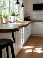 Modern white kitchen interior with live edge wooden countertop, subway tile backsplash, black pendant lights and potted herbs on windowsill in natural sunlight.