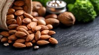 Raw almonds spilling from burlap sack onto dark stone surface with walnuts and fresh herbs in background, macro food photography for healthy eating concept.