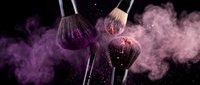 Makeup brushes with purple powder explosion on black background for cosmetic and beauty industry advertising campaigns.