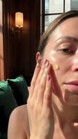 Young Caucasian woman applying facial cream in sunlit room with green velvet sofa. Morning skincare routine, anti-aging moisturizer application at home.