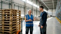 Mature Caucasian female warehouse worker in blue vest discussing logistics with young male supervisor in modern distribution center with wooden pallets.
