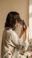 Woman with wet curly hair combing through tangles in natural sunlight wearing linen shirt for morning haircare routine and self-care wellness lifestyle.