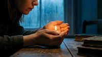 Young woman holding glowing light in cupped hands creating magical atmosphere in cozy indoor setting with books and window background for spiritual concepts.