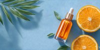 Orange vitamin C serum bottle with dropper surrounded by fresh orange slices and green leaves on blue background for skincare routine.