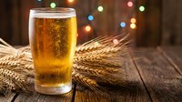 Golden beer glass with foam on rustic wooden table surrounded by wheat stalks and colorful bokeh lights creating warm festive atmosphere for celebrations.