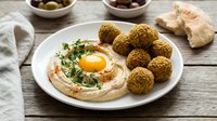 Crispy falafel balls served with creamy hummus topped with egg yolk and fresh herbs on a white plate, rustic wooden table.