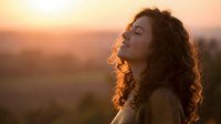 Young woman with curly hair enjoying peaceful sunset moment outdoors, eyes closed in meditation and relaxation against warm golden hour lighting.