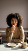 Young woman with curly afro hair and medium skin tone eating strawberry at table with coffee cup and cereal bowl, casual lifestyle portrait.