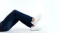 Young man legs in dark blue jeans and white sneakers sitting relaxed on white background. Casual fashion lifestyle concept for advertising and design.