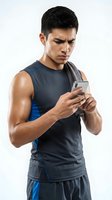 Young athletic man of East Asian descent wearing dark sleeveless sports shirt and blue shorts, holding smartphone while checking fitness app or workout data during training session against white background.