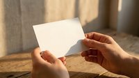 Hands holding blank white card with natural sunlight shadows creating warm atmosphere on wooden surface for mockup and presentation purposes.