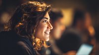 Profile view of young woman with curly hair smiling in warm ambient lighting, with blurred figures in background creating atmospheric depth.