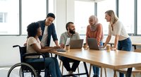 Diverse business team collaborating around laptop in modern office with natural light. Includes woman in wheelchair promoting workplace inclusion and accessibility.