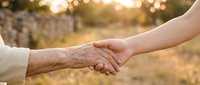 Young and elderly hands joining together in warm handshake outdoors during golden hour sunset, symbolizing intergenerational connection and support.
