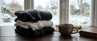 Stacked wool blankets with fringe next to ceramic mug and book on wooden table by window with winter landscape view.