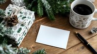 Festive holiday scene with blank card, coffee, and wrapped gift on wooden table surrounded by pine branches and decorative elements.