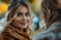 Young blonde woman with warm smile wearing cozy knit scarf having friendly conversation outdoors in autumn setting with soft bokeh background.