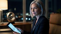 Mature woman with gray hair and light skin tone, wearing dark business blazer, holding tablet device in modern office at night with city lights visible through windows, professional corporate environment.