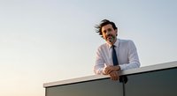 Caucasian male professional in business attire leaning on rooftop railing at sunset, wearing light blue shirt and dark tie, suitable for corporate and leadership concepts.