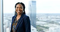 Young African woman in professional navy blazer smiling confidently at high-rise office window with cityscape view, corporate business portrait.
