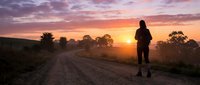 Runner jogging on rural dirt road at sunrise with golden sky and trees silhouetted in background, suitable for fitness and wellness content.