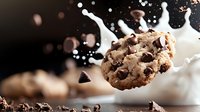 Chocolate chip cookie splashing into milk with dynamic white droplets against dark background, creating dramatic food photography with motion capture effect.