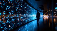 Silhouette of person observing interactive digital wall display with glowing blue light particles, creating futuristic atmosphere in modern exhibition space at night.