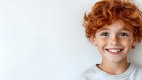Young child with bright red curly hair and genuine smile wearing gray t-shirt against white background, expressing joy and positive emotions.