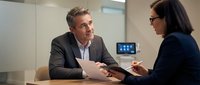 Middle-aged Caucasian male professional in business suit discussing documents with female colleague in modern office meeting room setting.