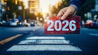 Hand holding red card with number 2026 over pedestrian crossing at sunset with urban street background and bokeh lights creating futuristic city mood.