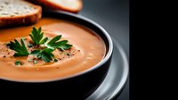 Creamy orange tomato soup garnished with fresh parsley and black pepper in dark ceramic bowl, served with bread slice on dramatic black background.